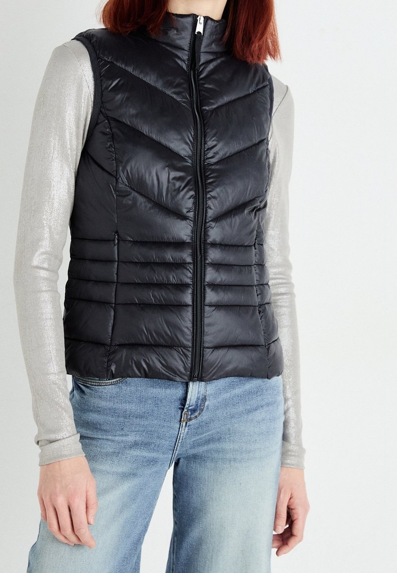 Gilet rembourré noir avec des panneaux en piqué vertical et en chevron, fermeture éclair et col montant. Porté par-dessus une chemise argentée à manches longues.