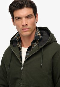 Oliwkowa kurtka z kapturem, zapinana na zamek, nałożona na niebieską i brązową koszulę w kratę, z pikowaną czarną podszewką i sznurkami do regulacji.