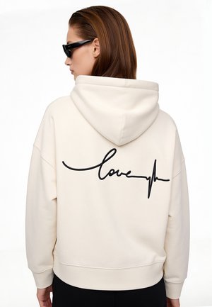 Vrouw met bruin haar die zwarte zonnebrillen draagt en een crèmekleurige hoodie met zwarte cursief "love" en een hartslagontwerp op de rug.