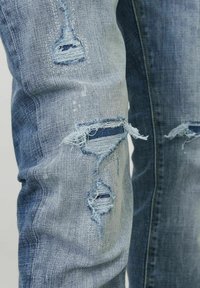 Jeans de denim desgastados de color azul claro con desgarros deshilachados en las rodillas, con un forro de color azul oscuro visible y una textura suave.