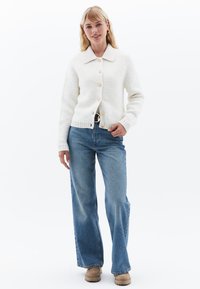 Vit, strukturerad cardigan med krage och knappstängning, i kombination med ljusblå vida jeans och bruna skor. En enkel, avslappnad design.