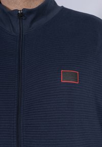 Navy zip-up jacket in textured fabric, featuring a high collar and a rectangular logo patch with red accents on the chest.