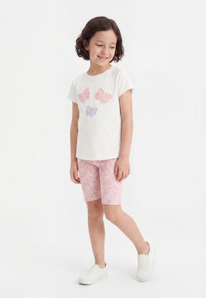 Pige iført hvid t-shirt med pink og blå sommerfugleprint, pink blomstrede shorts og hvide slip-on sko, smilende og kiggede ned.