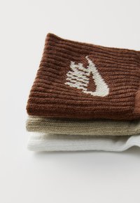 Rudos, smėlio ir baltos spalvos megztos kojinės, sudėtos viena ant kitos. Ruda kojinė turi baltą logotipą su tekstūruotomis linijomis. Minkšta, gumbuota medžiaga.
