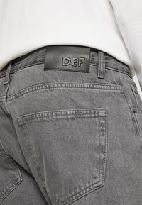 Vaqueros de mezclilla gris con un acabado texturizado. Presenta una etiqueta de cuero negro marcada "DEF" en la cintura. Incluye bolsillos traseros y costuras cosidas.