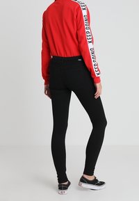 Sudadera roja con el texto "SIGUE ADELANTE" en las mangas, combinada con jeans skinny negros y zapatillas negras. Texturas suaves, ajuste casual.