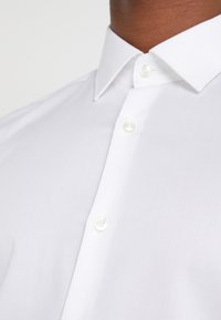 Camisa blanca de vestir con cuello puntiagudo, diseño de botones en la parte delantera y tejido texturizado. Detalles incluyen botones redondos blancos en la parte frontal.