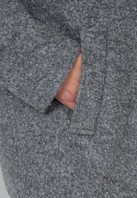 Manteau gris en tissu texturé avec un tissu tissé confortable. Présente une poche visible et un ourlet légèrement effiloché le long des manches.