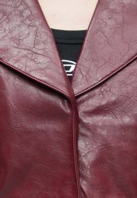 Burgunderfarbener Lederblazer mit strukturierter Oberfläche, versehen mit einem Reverskragen und einem leicht zerknitterten Aussehen. Schwarzes Oberteil darunter sichtbar.