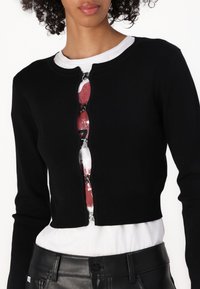 Schwarze, verkürzte Strickjacke mit rundem Ausschnitt, verziert mit dekorativen Knöpfen an der Vorderseite und in Schichten unter einem weißen T-Shirt.