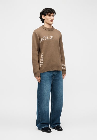 Νεαρός άνδρας με σγουρά μαλλιά, που φοράει καφέ πουλόβερ με λευκό κείμενο και φαρδιά μπλε τζιν, στέκεται μπροστά σε απλό ανοιχτό φόντο.