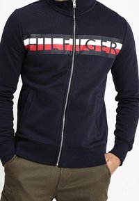 Giacca blu navy con zip e colletto alto. Presenta un logo orizzontale audace in rosso, bianco e nero sul petto. Polsini e orlo a coste.