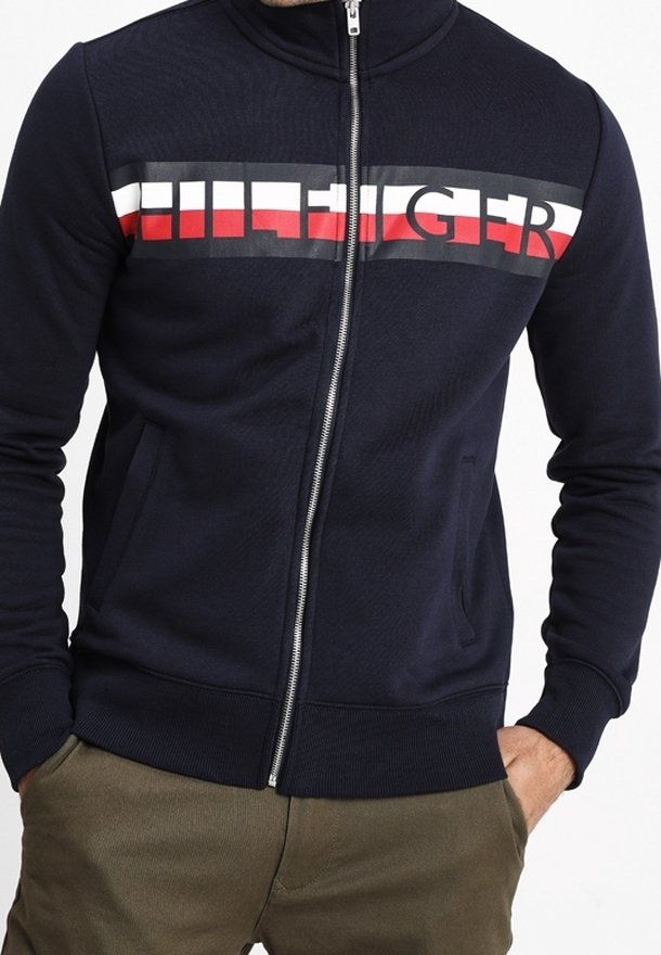 Giacca blu navy con zip e colletto alto. Presenta un logo orizzontale audace in rosso, bianco e nero sul petto. Polsini e orlo a coste.