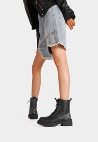 Stivali alla caviglia in pelle nera con punta rotonda, lacci frontali, suola in gomma spessa e dettagli decorativi sui lati.