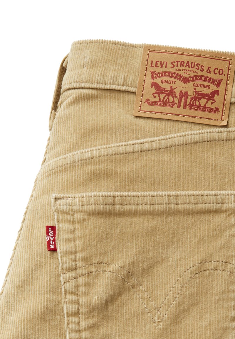 Pantalons en velours côtelé beige avec un patch en cuir griffé, une étiquette latérale rouge et un motif de couture courbé sur les poches arrière. Tissu texturé.