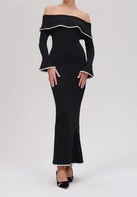 Vrouw in een lange zwarte off-shoulder aansluitende jurk met witte bies en uitlopende mouwen, gecombineerd met zwarte open teen hakken, op een effen achtergrond.