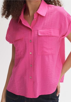 Frau trägt ein knallpinkes kurzärmeliges Hemd mit zwei Brusttaschen und dunkle Hosen, Hand auf der Hüfte ruhend.