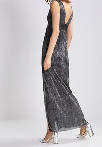 Full-length black dress with a pleated design and a low back. Features silver accents and a fitted waist, paired with metallic block-heel shoes.