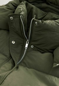 Geacă puf dintr-o nuanță de verde închis, cu un fermoar metalic, nasturi snap de culoare argintie și un căptușel interior texturat. Materialul pare neted și are un design matlasat.
