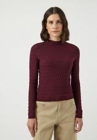 Bordowy sweter z długim rękawem z teksturowanym wzorem w kwadratowe motywy i wysokim kołnierzem, zestawiony z beżowymi spodniami.