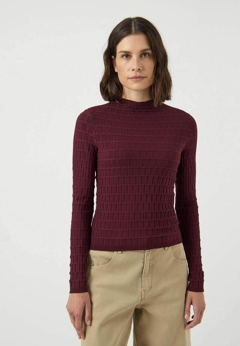 Bordowy sweter z długim rękawem z teksturowanym wzorem w kwadratowe motywy i wysokim kołnierzem, zestawiony z beżowymi spodniami.