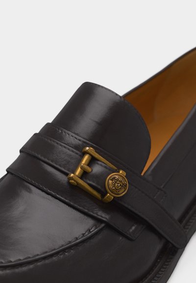 Κοντινό πλάνο μαύρου δερμάτινου μοκασίνι με χρυσή πόρπη που έχει έμβλημα κεφαλής Μέδουσας στο λουρί, με εμφανείς λεπτομερείς ραφές και ομαλή επιφάνεια.