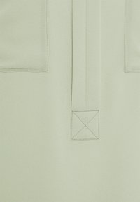 Blusa verde chiaro con una texture liscia, una patta con bottoni e due tasche frontali con dettagli quadrati sul fondo.