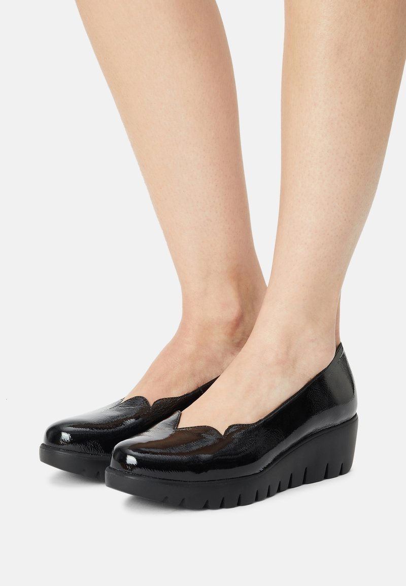 Chaussures compensées en cuir verni noir avec bords festonnés portées pieds nus, vues de côté devant sur un fond blanc.