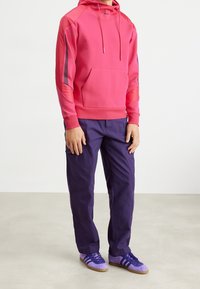 Hoodie w jaskrawym różu z przednią kieszenią kangurką i kontrastującymi paskami po bokach. W połączeniu z ciemnofioletowymi spodniami cargo i fioletowymi sneakersami.