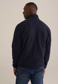 Tumesinine fliiskindel jakker kõrge kraega, pikkade varrukatega ja sirge lõikega. Omab siledat tekstuuri ja puhta disaini.