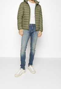 Chaqueta acolchada color verde oliva con costuras horizontales, camiseta blanca, vaqueros ajustados azules y zapatillas blancas con detalles en gris claro.