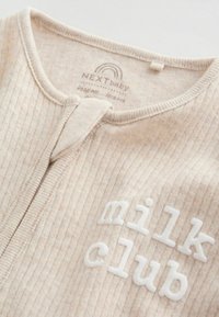 Zbliżenie na beżowy prążkowany niemowlęcy body z przednim zamkiem błyskawicznym i białym, fakturowanym napisem "milk club" na piersi.