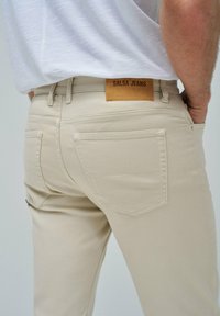 Jeans salsa beige con parche de cuero, diseño de cinco bolsillos y detalles de costura visibles. Material de algodón suave.