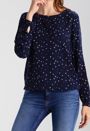 Frau trägt eine dunkelblaue Langarmbluse mit kleinen, mehrfarbigen Blumenmustern und blaue Denim-Jeans.