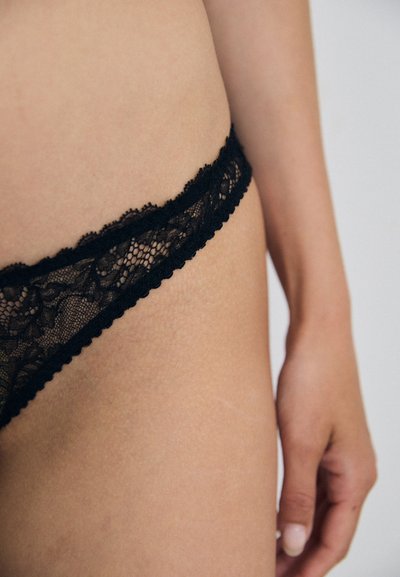 Black lace thong featuring a scalloped edge and floral pattern, with soft, sheer texture and minimal coverage, displayed against skin.