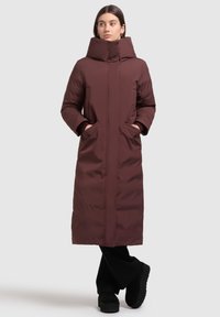 Burgundowa długa kurtka puchowa z wysokim kołnierzem, bocznymi kieszeniami i gładką teksturą. Noszona z czarnymi spodniami dzwonami i butami na platformie.