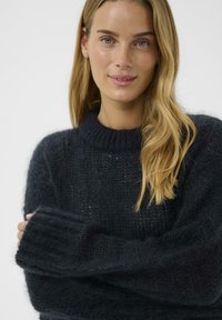 Tumša, teksturēta džemperis ar ribotu augstu apkakli un garām piedurknēm, piemīt vaļīga piegriezuma un mīksta adījuma materiāla.