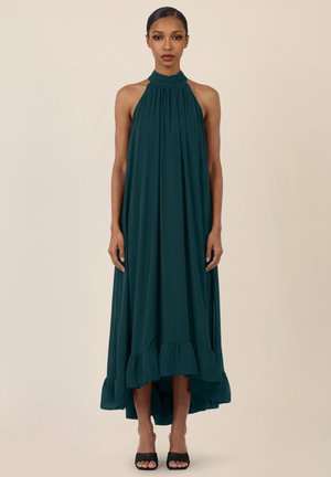 Kobieta ubrana w ciemnozieloną, bezrękawnikową sukienkę z wysokim dekoltem na halter z falbaną u dołu oraz czarne sandały na obcasie z odkrytymi palcami, stojąca na tle jednolitego beżowego tła.