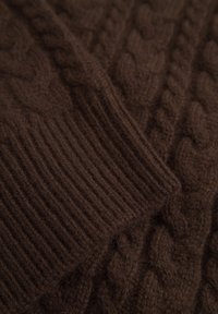 Dettaglio di un maglione di colore marrone lavorato a maglia con un motivo a treccia e polsini a coste. Il materiale sembra morbido e texturizzato.