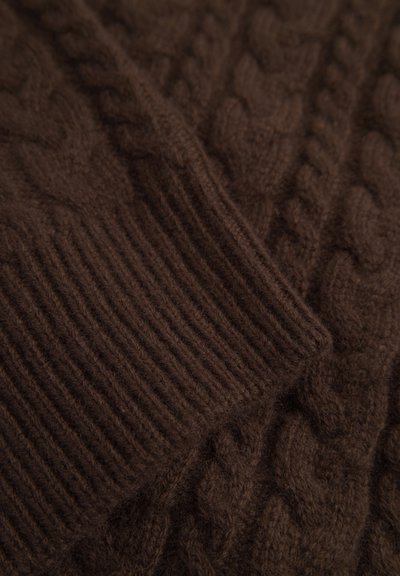 Rjav pleteni pulover z detajli, ki vključujejo pleten vzorec in rebraste manšete. Material deluje mehak in teksturiran.