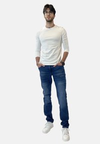 Camisa blanca de manga larga, jeans azules y zapatillas blancas. Reloj de pulsera notable y gafas. Corte recto con ligero estiramiento en el denim.