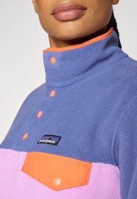 Fleece pullover in purple and pink with orange accents. Features a high collar, snap buttons, and a front pocket with branding.