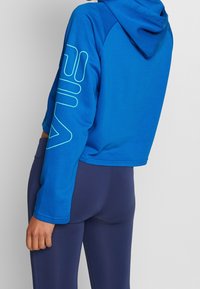 Person som bär en blå kortärmad huvtröja med en stor ärm logotyp och marinblå tajta leggings, som står mot en enkel ljus bakgrund.