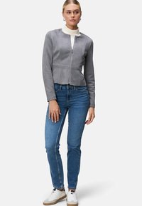 Giacca corta in finta suede grigia con zip frontale, maniche lunghe e cuciture. Indossata sopra un dolcevita color crema, abbinata a jeans blu e sneakers bianche.
