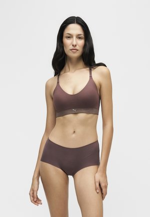 Femme aux longs cheveux foncés portant un soutien-gorge de sport marron assorti et un sous-vêtement taille haute, debout devant un fond clair uni.