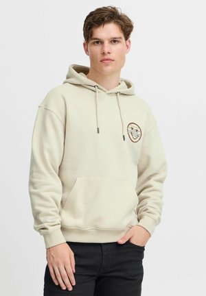 Beige hoodie with a front pocket, drawstring hood, and a round patch featuring printed text and designs. Cotton blend fabric, relaxed fit.