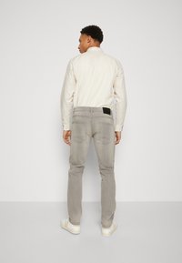 Gray stretchy jeans with a straight leg fit, back pockets, and a black leather logo patch, paired with a light-colored button-up shirt.