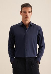 Camicia a maniche lunghe blu scuro realizzata in tessuto morbido con colletto abbottonato e bottoni abbinati. Indossata con pantaloni neri.