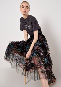 Czarna tiulowa spódnica z kwiatowym wzorem, warstwowa z falbanami, noszona na szarym t-shircie z grafiką. Spódnica ma wielokolorowe kwiatowe motywy i przezroczystą teksturę.