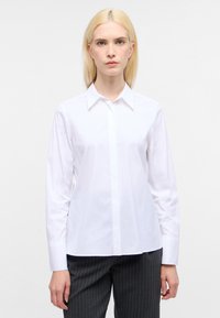 Weißes Hemd mit klassischem Kragen, langen Ärmeln und einer glatten, knackigen Textur. Kombiniert mit schwarzen Nadelstreifen-Hosen.